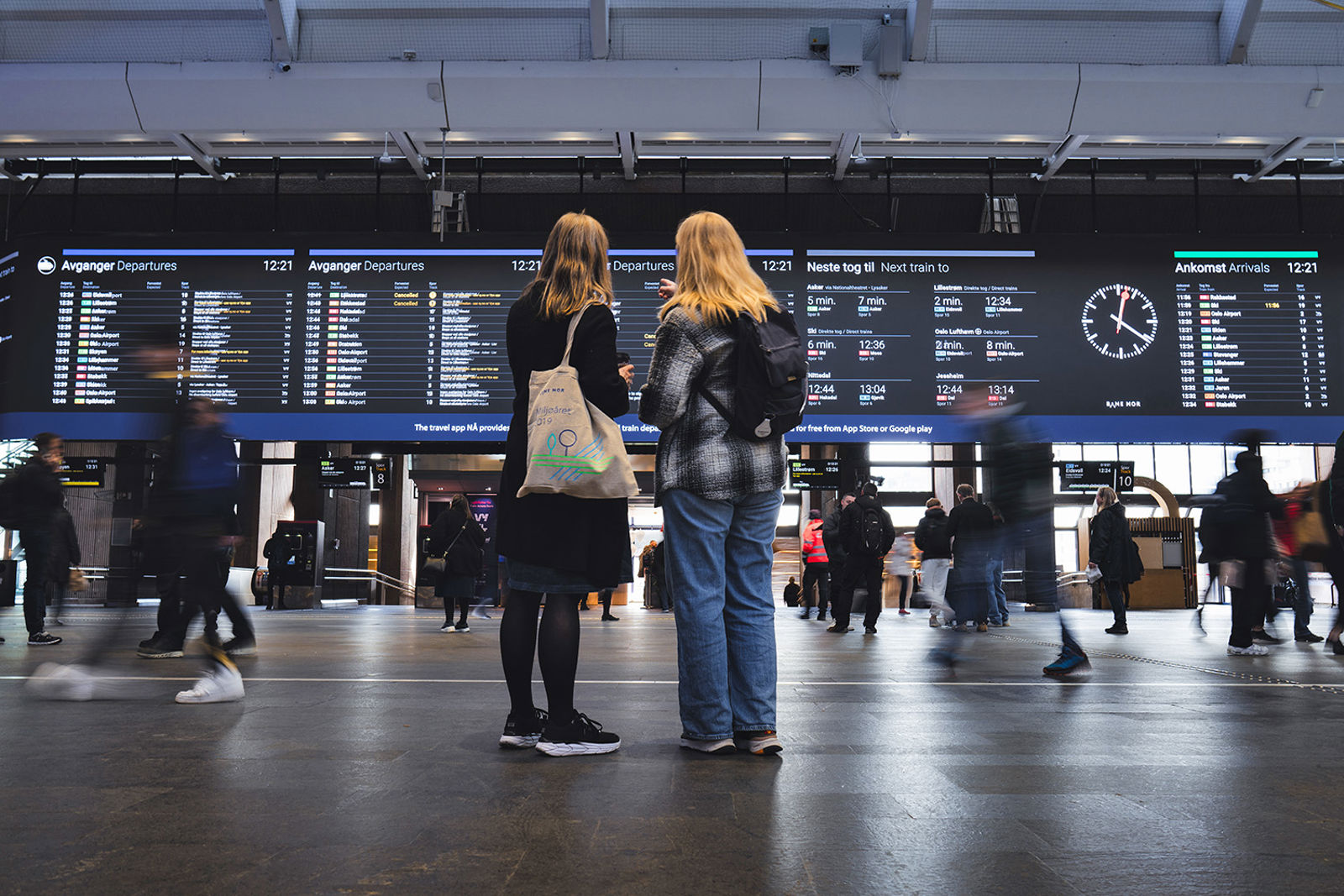 To personer står med ryggen til og ser på den store informasjonstavlen på Oslo S.