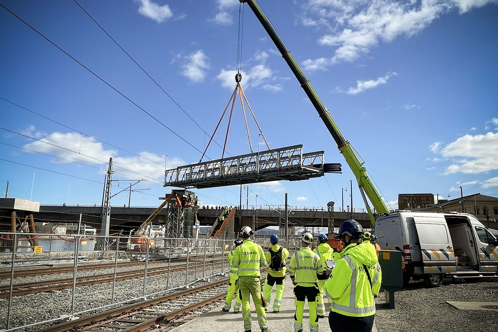 Flere gulkledde personer står langs jernbanesporet og ser på et bruelement som blir løftet på plass.