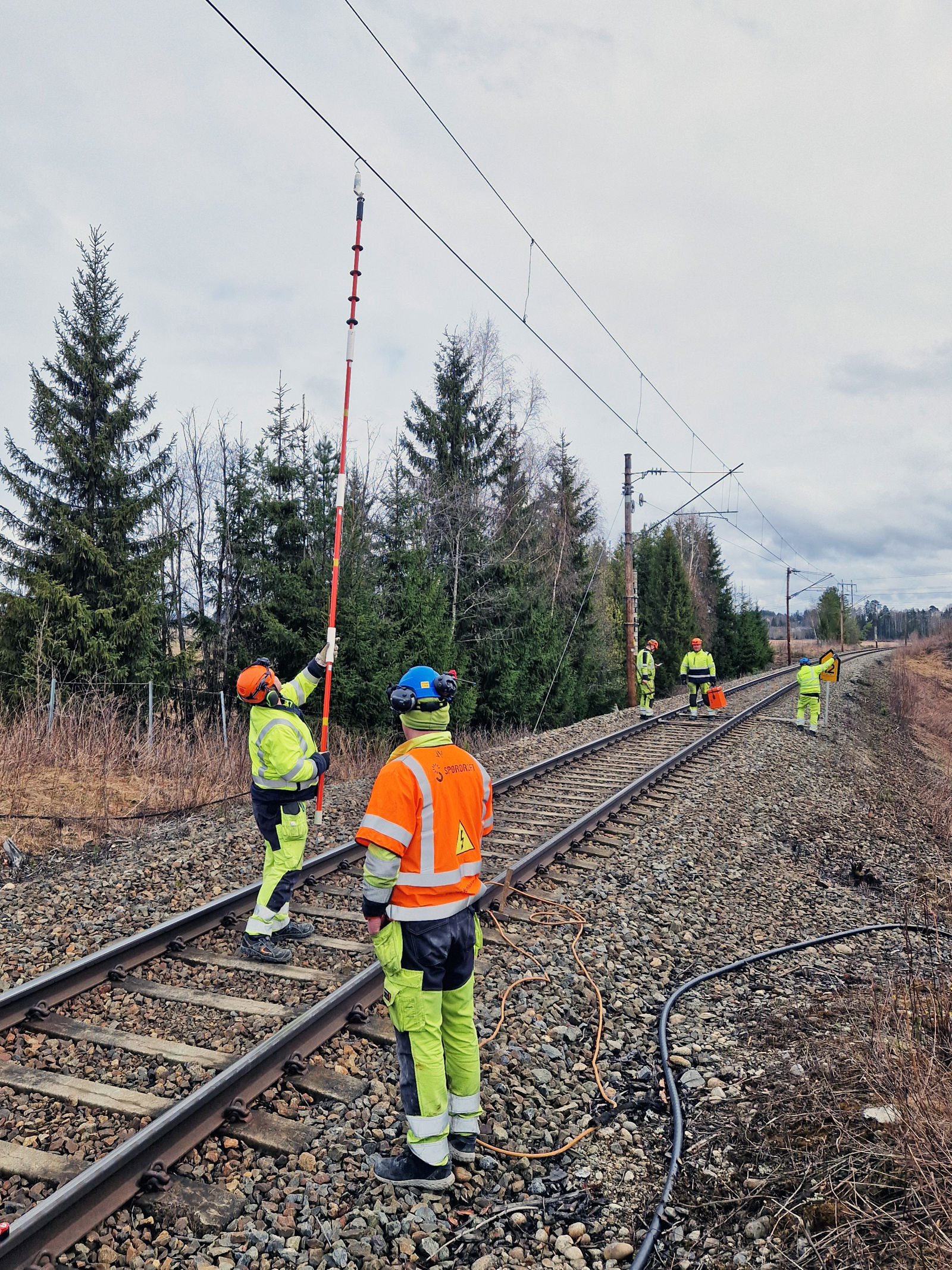 To mann i vernetøy henger en stang med krok i kontaktledningen som går over skinnegangen.
