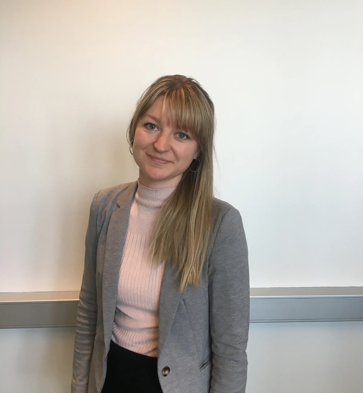 Portrettbilde av Nina Valle med rosa genser, grå blazer, langt hår og pannelugg. 