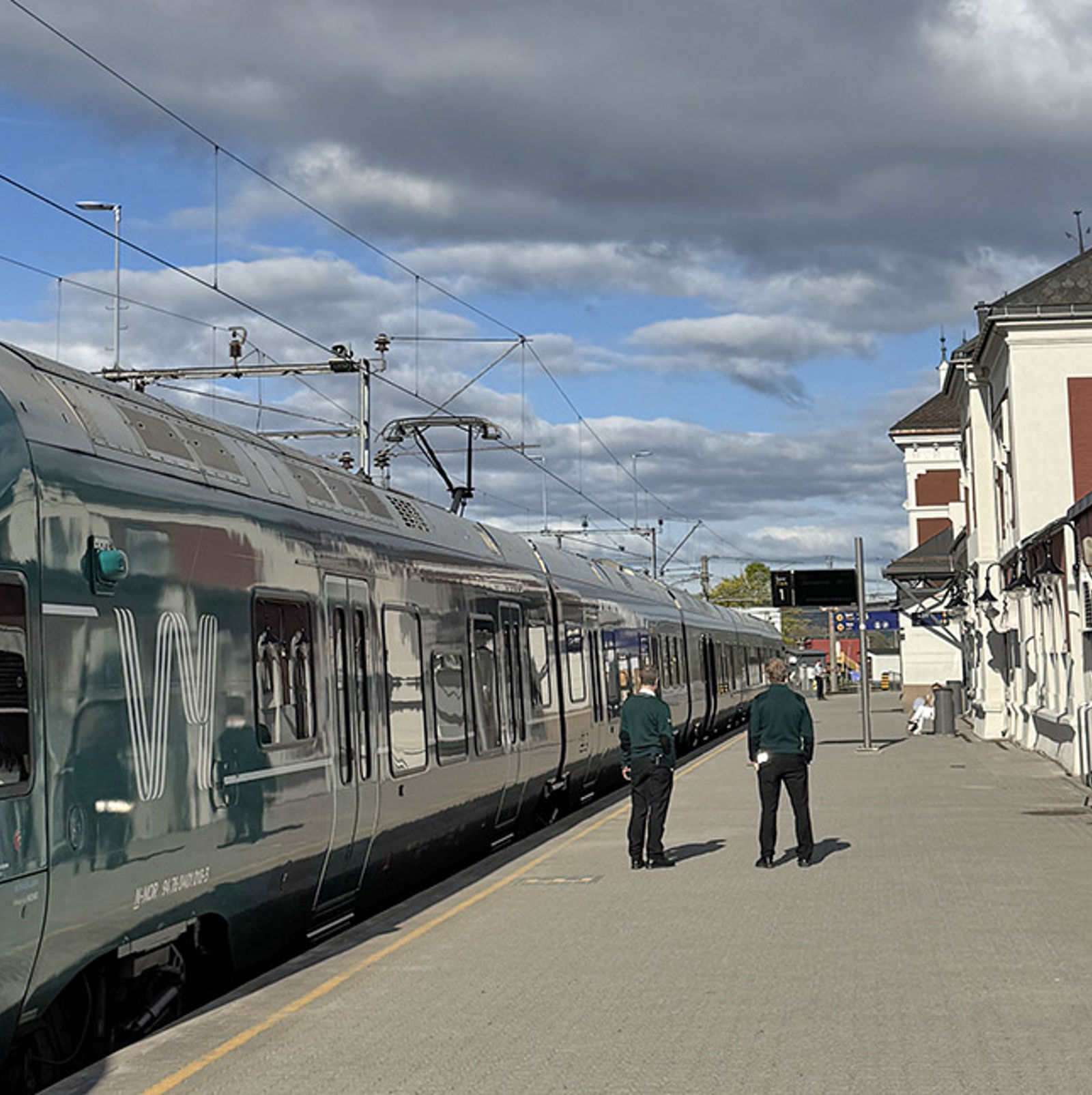 Grønt Vy-tog til venstre, personer på plattformen, stasjonsbygningen og sykkelparkering til høyre.