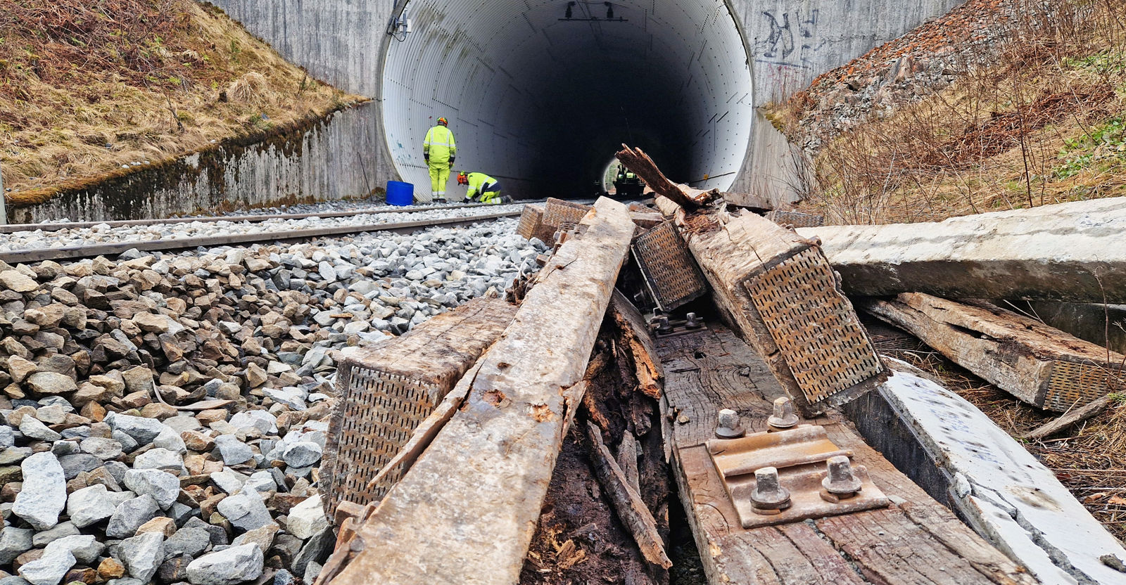 Noen gamle, råtne tresviller i forgrunn, jernbanespor som går inn i en tunnel i bakgrunn. 