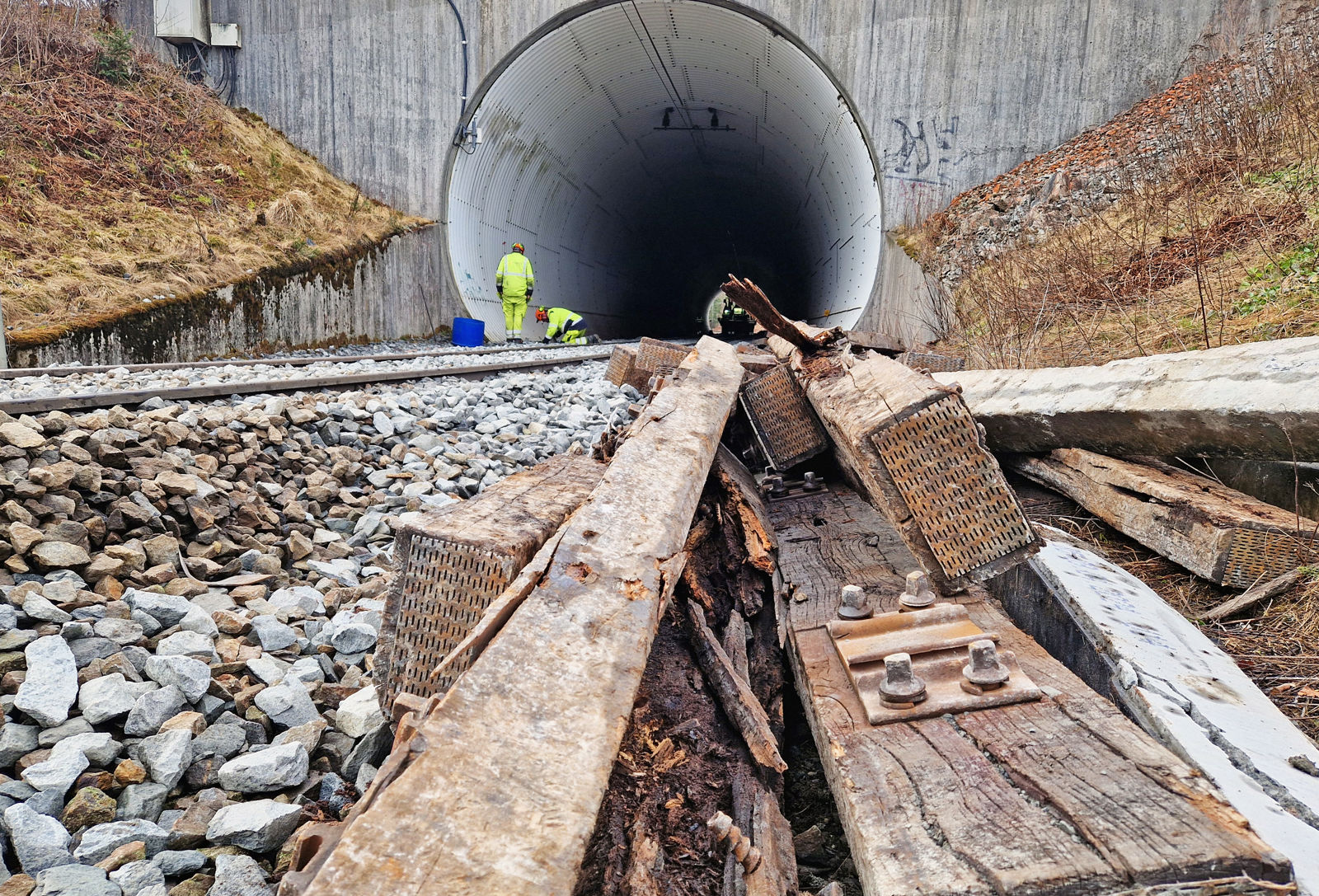 Noen gamle, råtne tresviller i forgrunn, jernbanespor som går inn i en tunnel i bakgrunn.