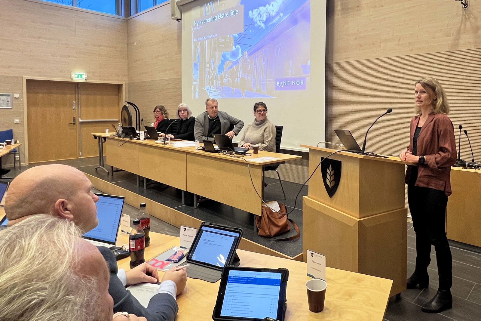 Kvinne står og prater i mikrofon foran en sal med menn og kvinner.