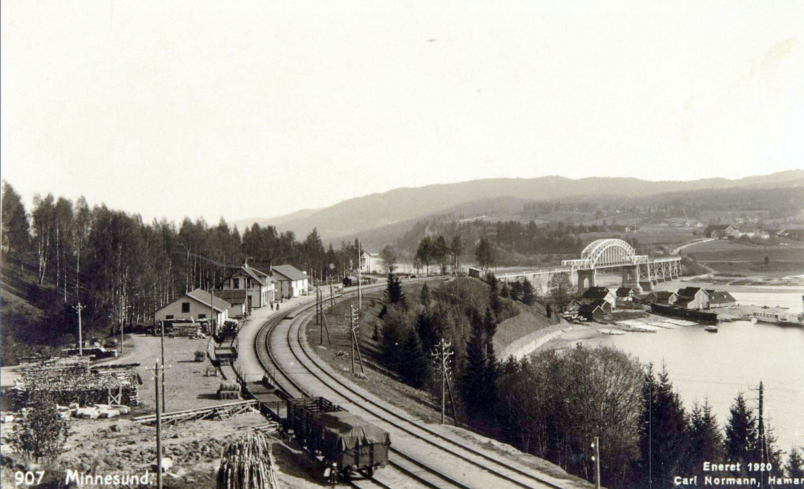 Stasjonen med godsvogner i forgrunnen, to personer på plattformen, Minnesund i bakgrunnen.
