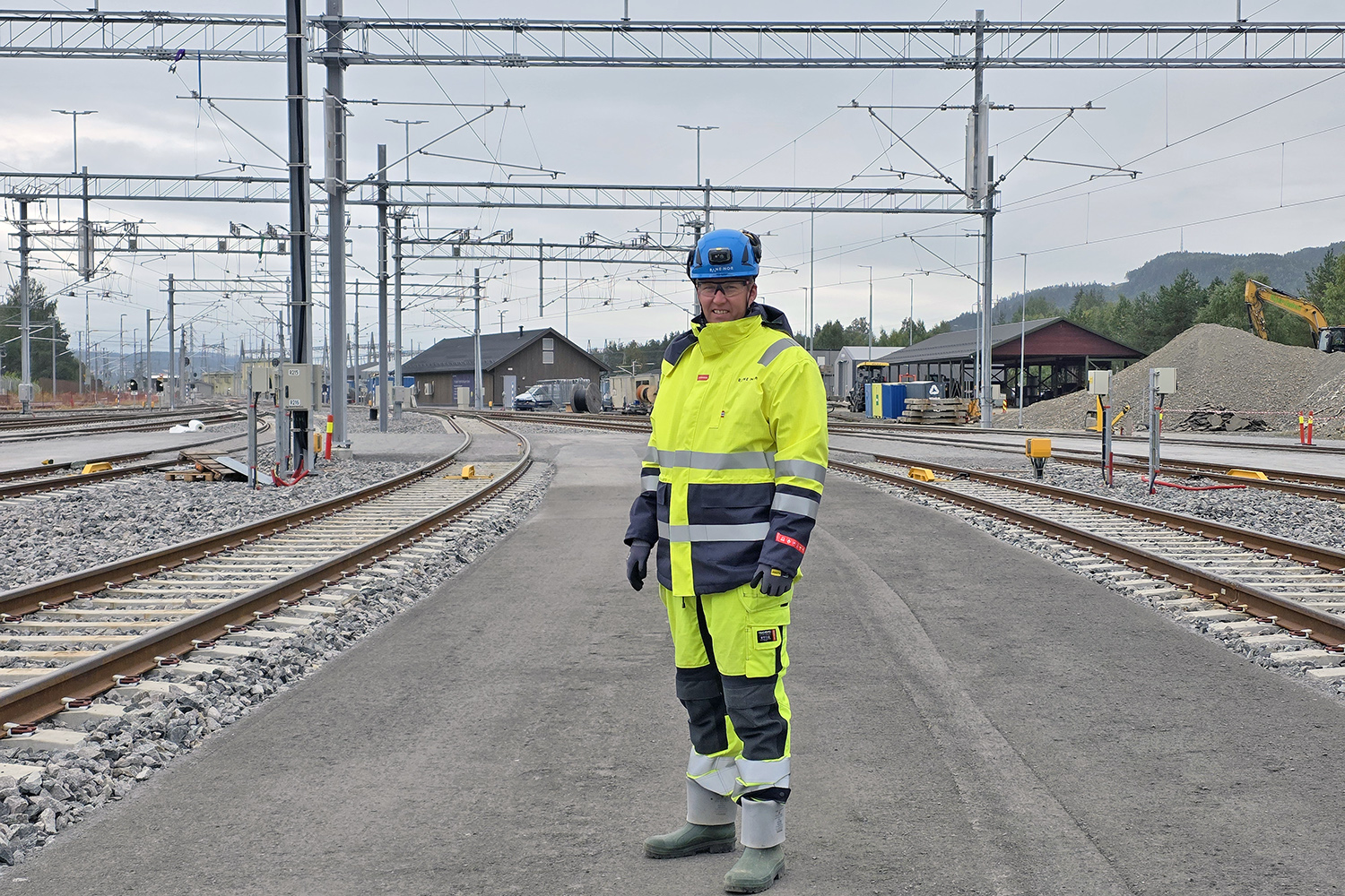 Prosjektlederen iført vernetøy og hjelm med spor og bygninger i bakgrunnen.