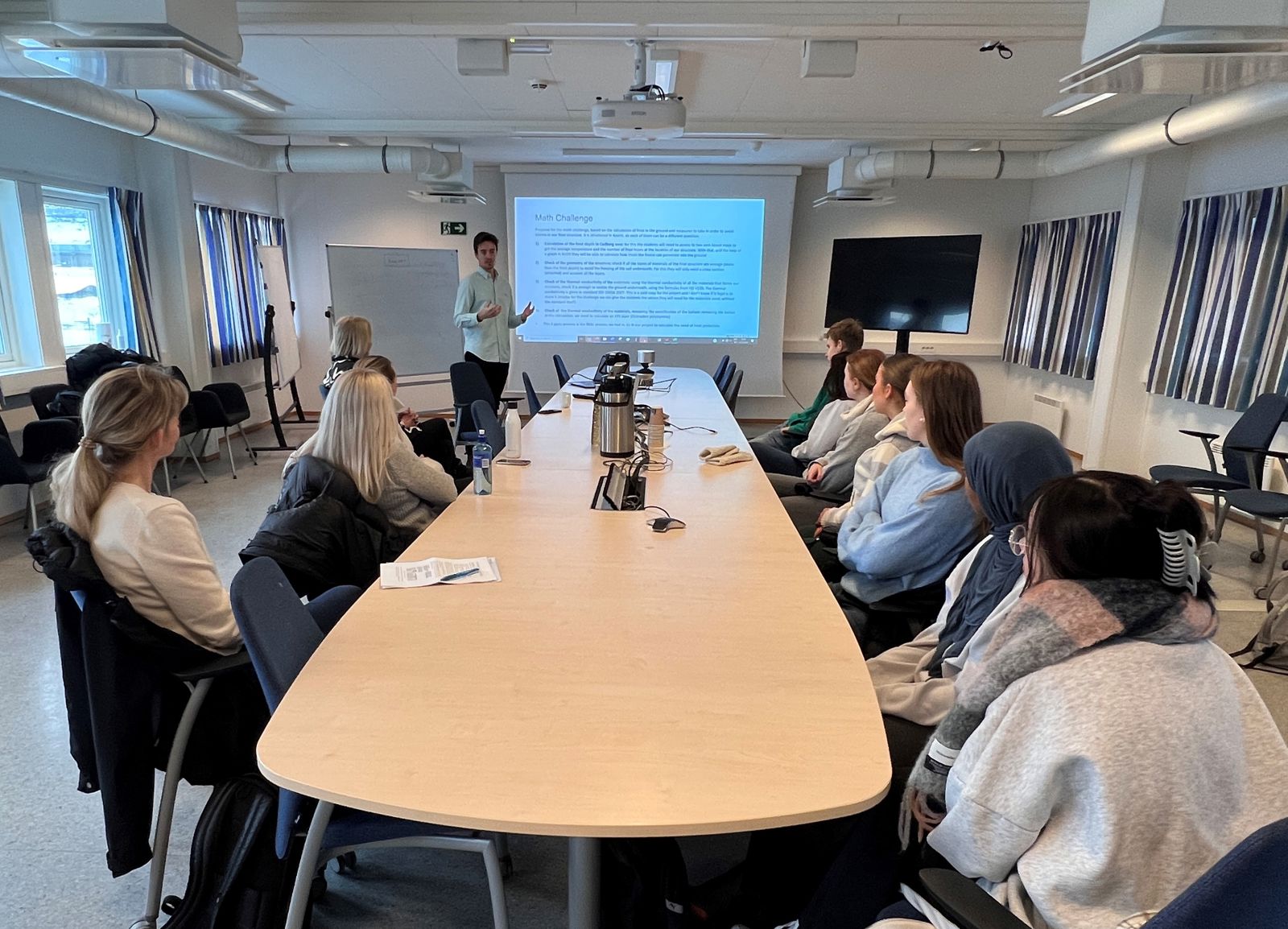 Ungdom sitter rundt et bord og helt fremst står en mann og orienterer ved en PP-presentasjon